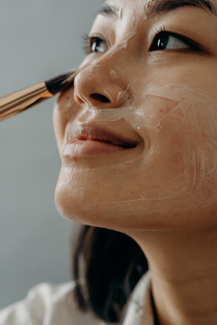 Close-up of an Asian woman applying face cream with a brush, promoting skin care and wellness.